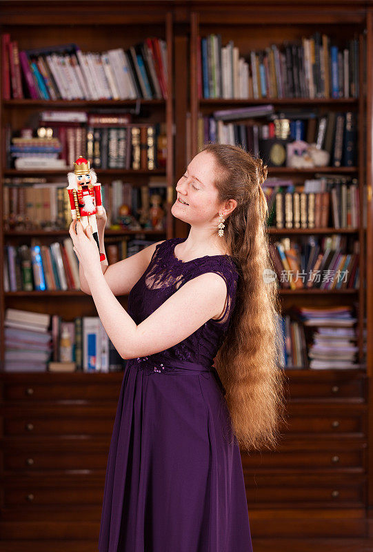 Young girl with long dark blonde hair in a purple dress stands on New Year's eve admiring a Nutcracker figurine gift.